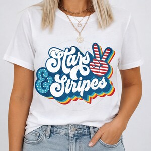 Peut inclure: T-shirt blanc avec l'inscription "Stars & Stripes" dans une police rétro et un signe de paix. Le motif utilise les couleurs rouge, blanc et bleu, avec des éléments d'étoiles et de rayures.