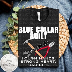 Könnte beinhalten: Dunkelgraues T-Shirt mit dem Text "BLUE COLLAR BUILT" und "TOUGH HANDS, STRONG HEART, DAD LIFE". Das Design beinhaltet Illustrationen von Hammer, Schraubenschlüssel und Zange. Ein rundes Logo mit dem Text "Comfy Cozy Things SHIRT STORE" befindet sich unten links.