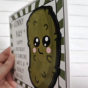 Peut inclure: Une carte blanche avec des rayures vertes et blanches sur le c&ocirc;t&eacute;. La carte porte le texte "Joyeux anniversaire &agrave; quelqu'un qui est un tr&egrave;s gros cornichon..." et un cornichon de dessin anim&eacute; avec un visage. Le cornichon est vert avec un contour noir et a un grand sourire et de grands yeux.