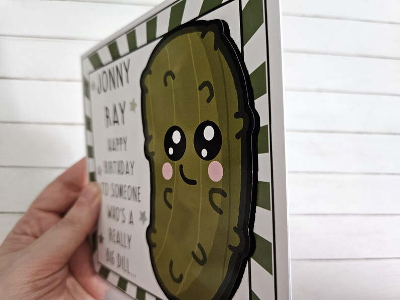 Peut inclure: Une carte blanche avec des rayures vertes et blanches sur le c&ocirc;t&eacute;. La carte porte le texte "Joyeux anniversaire &agrave; quelqu'un qui est un tr&egrave;s gros cornichon..." et un cornichon de dessin anim&eacute; avec un visage. Le cornichon est vert avec un contour noir et a un grand sourire et de grands yeux.