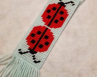 Może przedstawiać: Ręcznie robiona zakładka do książki z jasnoniebieskim tłem i dwoma czerwonymi biedronkami. Każda biedronka ma czarne kropki i czarny kontur. Zakładka ma frędzle na obu końcach. Uroczy dodatek dla miłośników książek.