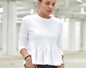 Puede incluir: Una mujer con una blusa blanca con volantes y pantalones grises holgados. Está de pie en un entorno industrial blanco.