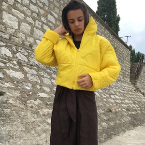 May include: A cropped, bright yellow puffer jacket with a high collar and a front zipper. The jacket is paired with dark brown, wide-leg pants. The outfit is worn outdoors against a stone wall background.