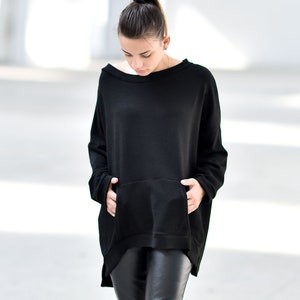 May include: A woman wearing a black oversized sweatshirt with a kangaroo pocket and black leather pants. She is standing on a concrete floor.