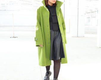 May include: A long, bright green coat with a high collar and button closure. The coat is paired with a black top, skirt, and patterned tights. Black shoes complete the outfit. The coat features a unique design on the sleeve.