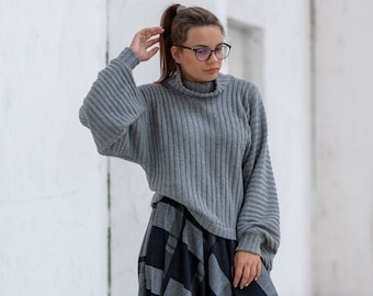 May include: A woman wearing a gray turtleneck sweater and a long black and gray plaid skirt. She is standing in front of a white wall.