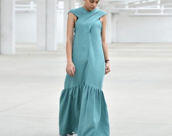 May include: A long, flowing teal dress with a unique criss-cross neckline and a ruffled hem. The dress is made of a soft, lightweight fabric and is worn with white sneakers. The setting is a large, open, white-walled space.