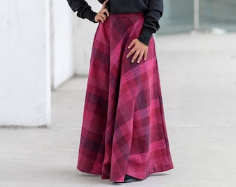 May include: A woman wearing a black mock neck sweater and a long, pink and black plaid skirt. She is standing in a white room with a concrete floor.