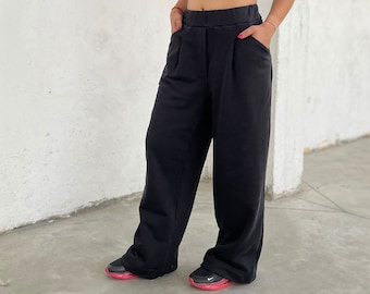 May include: A woman wearing a black turtleneck crop top and black wide-leg pants. She is wearing black glasses and has her hair pulled back.