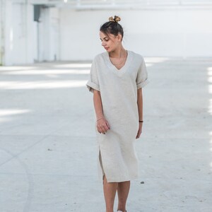May include: A woman wearing a light beige linen dress with a v-neck and short sleeves. She is wearing white sneakers and has her hair pulled back in a bun.