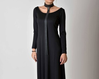 Puede incluir: Un vestido negro maxi con mangas largas y un escote redondo. El vestido presenta un largo y fino choker de cuero negro con un cierre plateado.