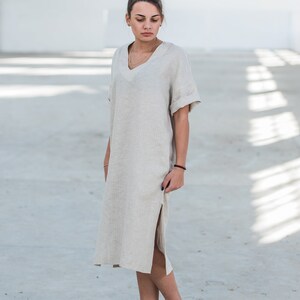 May include: A woman wearing a light beige linen dress with a side slit and short sleeves. She is wearing white sneakers and has her hair pulled back in a bun.