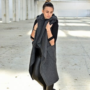 May include: A woman wearing a gray hooded vest over a black top and black pants. She is standing in a large, empty space with white pillars.