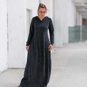 May include: A woman wearing a long, dark gray hooded dress with a relaxed fit. The dress has a slightly textured fabric and long sleeves. She is standing in a white hallway.