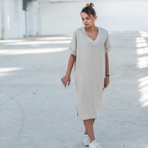 May include: A woman wearing a light beige linen dress with a v-neck and short sleeves. She is standing in a white industrial space and wearing white sneakers.