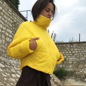 May include: A person wearing a bright yellow puffer jacket with a high collar and dark brown, wide-leg pants. The person is also wearing black sneakers with blue accents. The background features a stone wall and a cloudy sky.