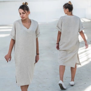 May include: A woman wearing a light beige linen dress with a v-neck and short sleeves. The dress has a side slit and is knee-length.
