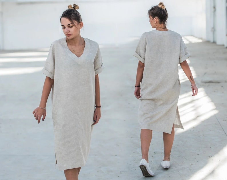May include: A woman wearing a light beige linen dress with a v-neck and short sleeves. The dress has a side slit and is knee-length.