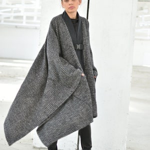 Pu&ograve; includere: Una donna indossa un lungo cappotto a quadri bianco e nero con un dolcevita nero sotto. &Egrave; in piedi in una stanza bianca con un muro bianco dietro di lei. Indossa pantaloni neri e scarpe nere.