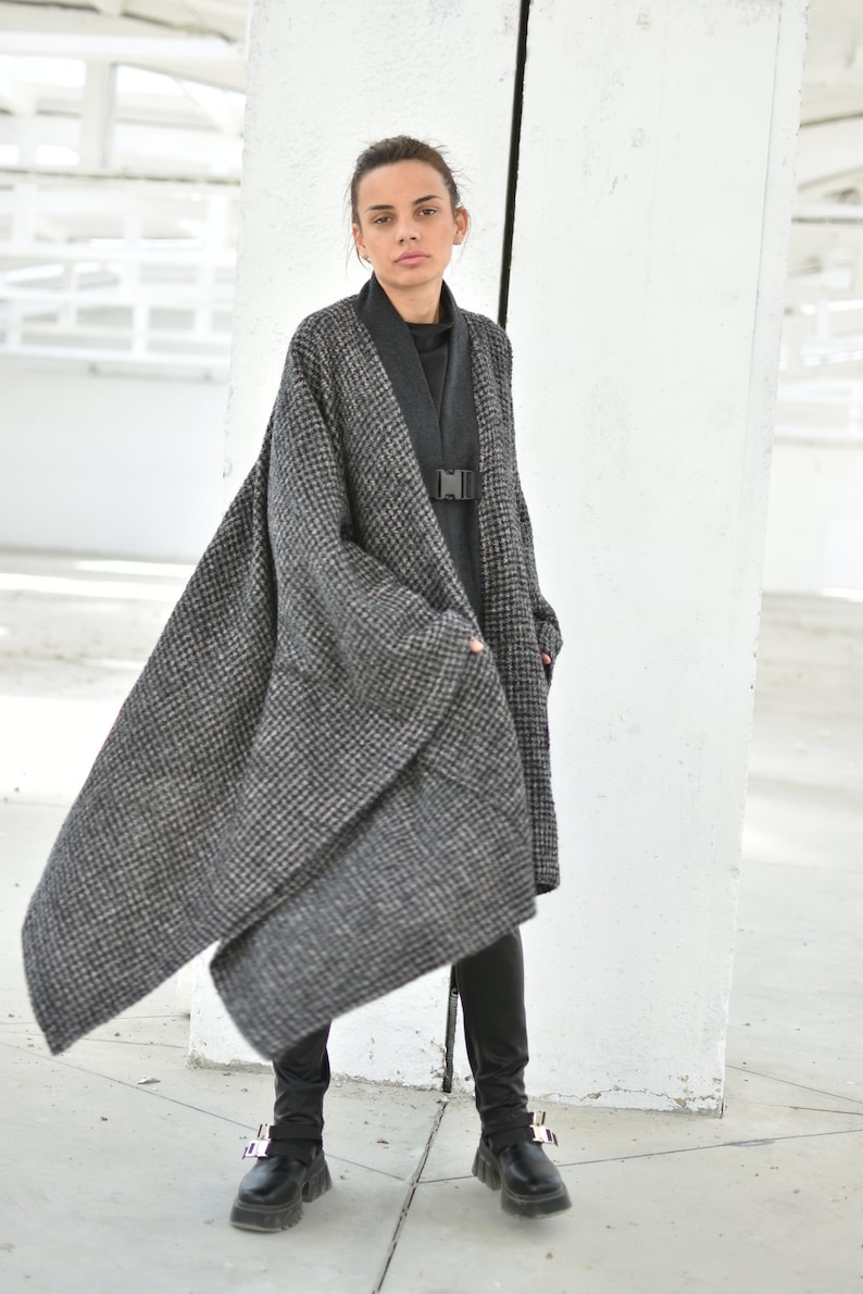 Pu&ograve; includere: Una donna indossa un lungo cappotto a quadri bianco e nero con un dolcevita nero sotto. &Egrave; in piedi in una stanza bianca con un muro bianco dietro di lei. Indossa pantaloni neri e scarpe nere.