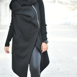 May include: A black hooded vest with an asymmetrical design and a gold zipper. The vest is layered over a black turtleneck and black leggings. A black watch is visible on the wrist. The outfit is all black.
