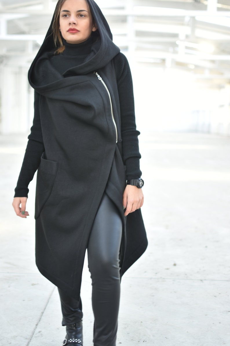 May include: A black hooded vest with an asymmetrical design and a gold zipper. The vest is layered over a black turtleneck and black leggings. A black watch is visible on the wrist. The outfit is all black.