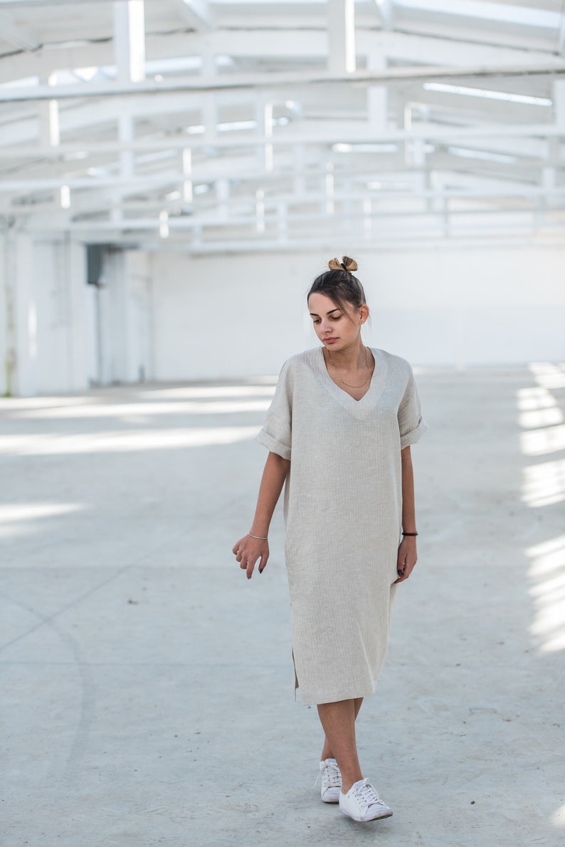 May include: A woman wearing a light beige linen dress with a v-neck and short sleeves. She is standing in a white industrial space and wearing white sneakers.