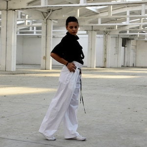 May include: A woman wearing a black blouse with a high neck and a white pair of wide-leg pants. She is standing in a large, empty warehouse with a white metal roof.