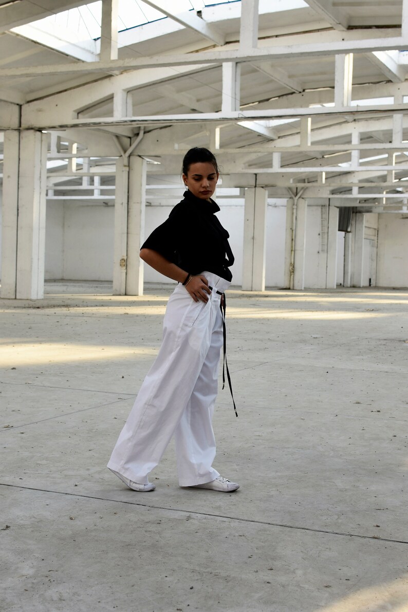 May include: A woman wearing a black blouse with a high neck and a white pair of wide-leg pants. She is standing in a large, empty warehouse with a white metal roof.