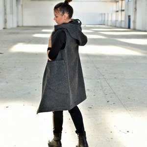 May include: A long, gray hooded vest with a relaxed fit. The vest is sleeveless and has a large hood. The model is wearing black pants and black boots.