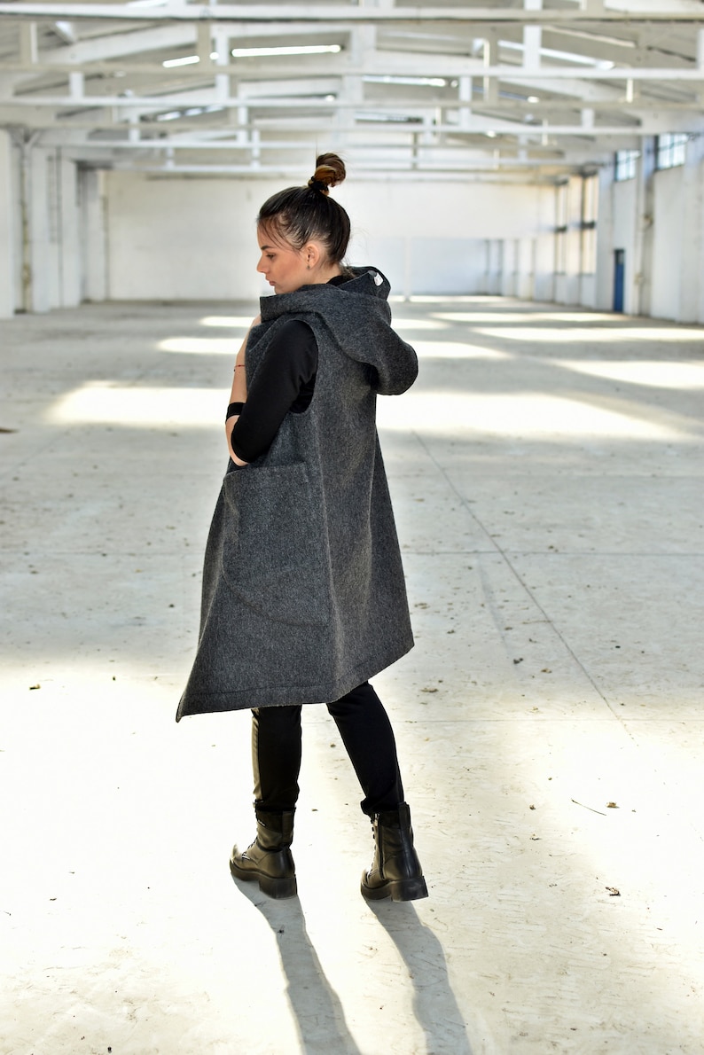May include: A long, gray hooded vest with a relaxed fit. The vest is sleeveless and has a large hood. The model is wearing black pants and black boots.