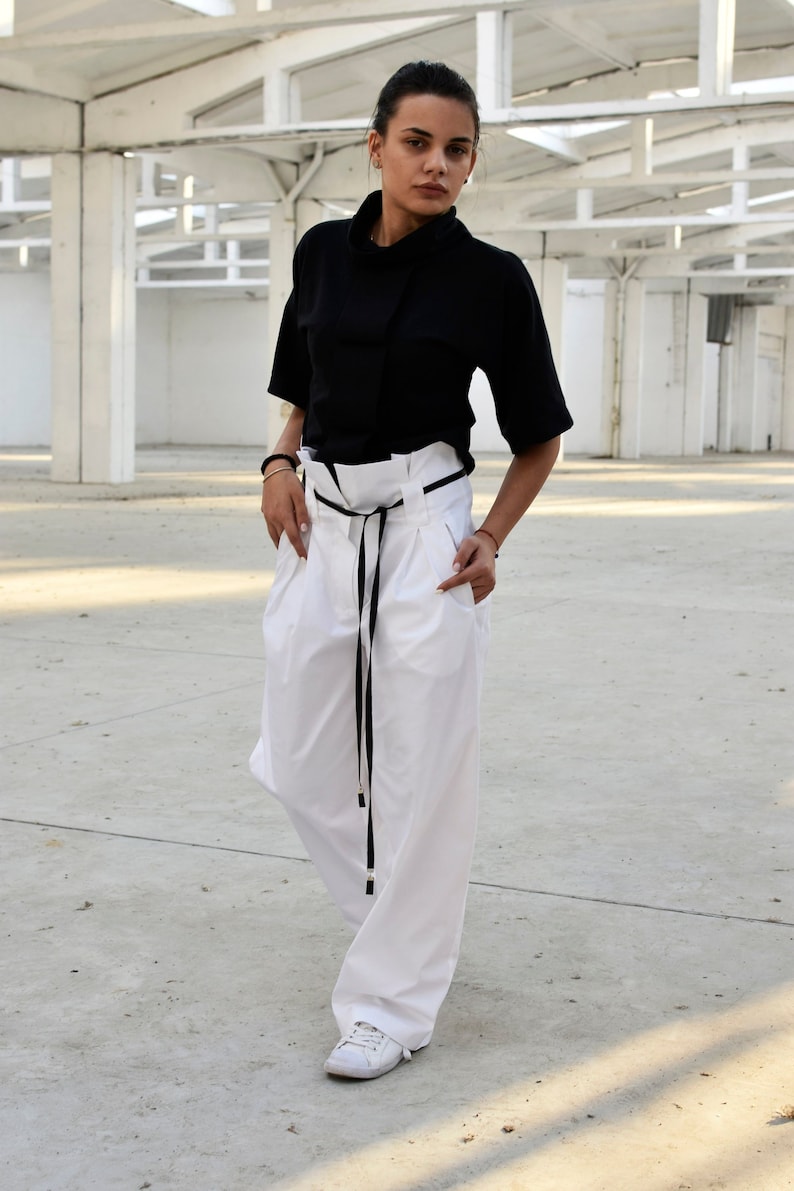 May include: A woman wearing a black short-sleeved shirt with a high neckline and white wide-leg pants with a black belt. She is standing in a white industrial setting.