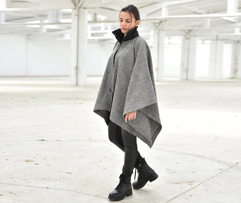 May include: A gray and black wool poncho with a high collar and a zipper closure. The poncho is worn by a person standing in a white room.