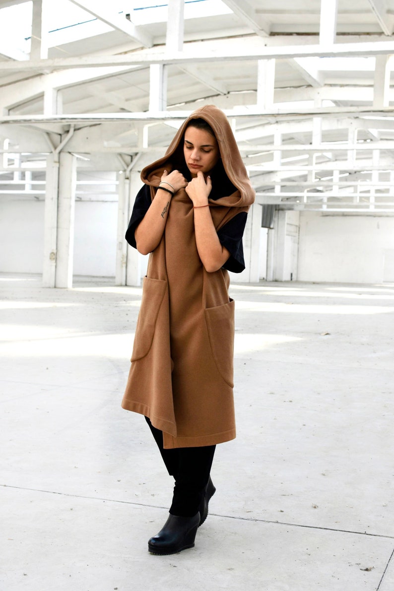 May include: A brown hooded vest with large pockets. The vest is worn over a black shirt and black trousers. The person is standing in a white industrial setting.