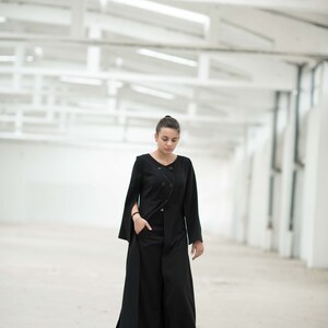 May include: A woman wearing a black long-sleeved jacket with a double-breasted closure and a long, flowing skirt. She is standing in a white, industrial-looking space.