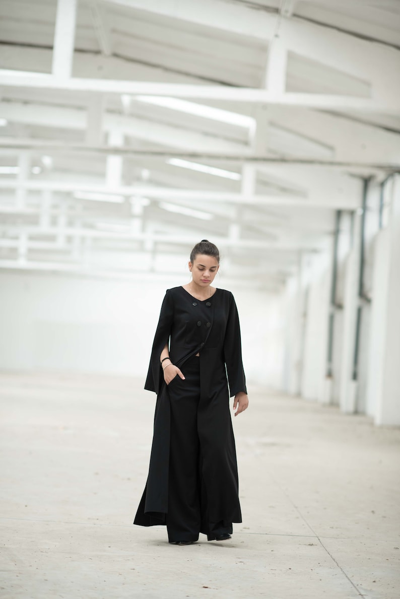 May include: A woman wearing a black long-sleeved jacket with a double-breasted closure and a long, flowing skirt. She is standing in a white, industrial-looking space.