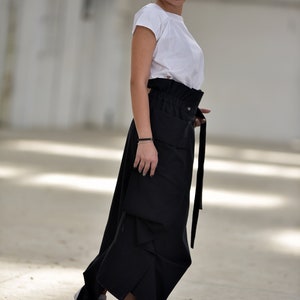 May include: A woman wearing a white t-shirt and a black, asymmetrical skirt with a high waist and a belt. She is wearing white sneakers.