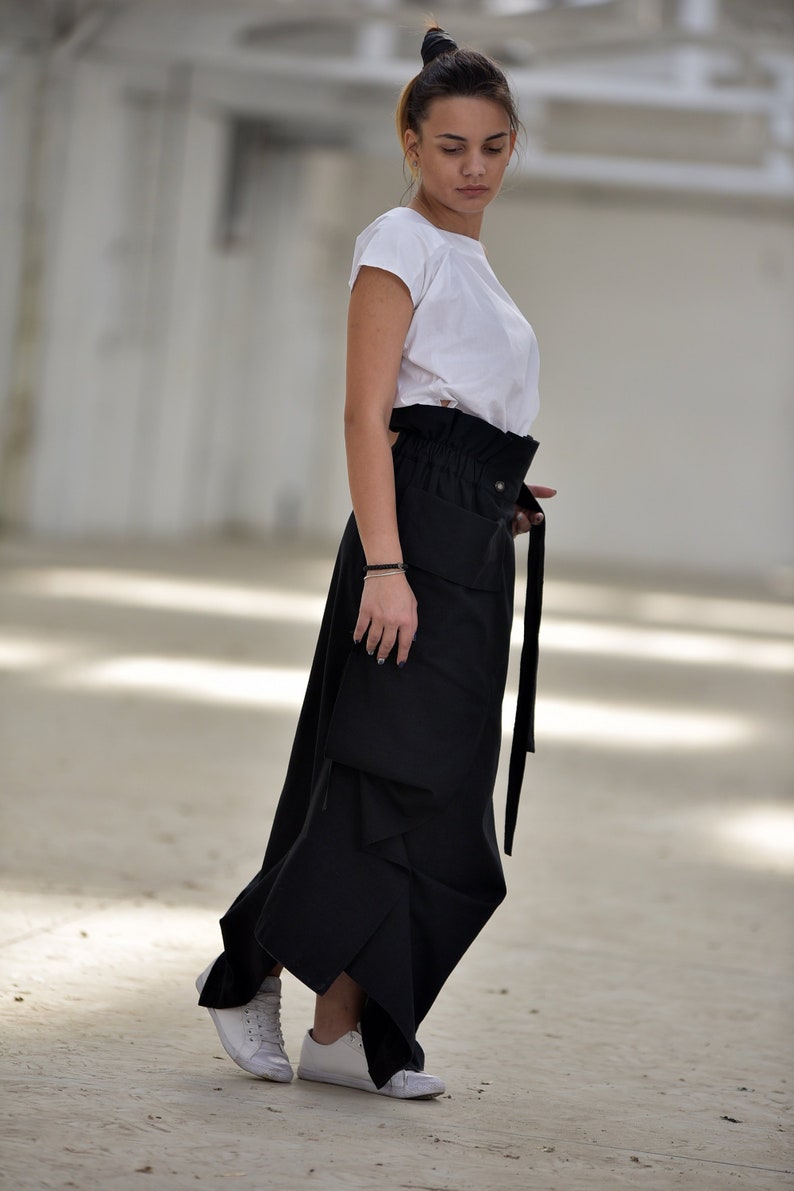 May include: A woman wearing a white t-shirt and a black, asymmetrical skirt with a high waist and a belt. She is wearing white sneakers.