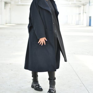 May include: A woman wearing a long, dark blue coat with a large collar and a black turtleneck shirt. She is wearing black leather boots with silver buckles.