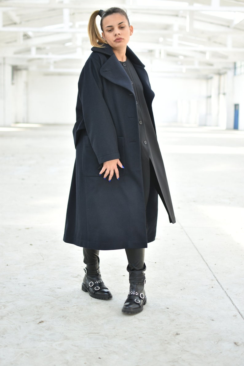 May include: A woman wearing a long, dark blue coat with a large collar and a black turtleneck shirt. She is wearing black leather boots with silver buckles.