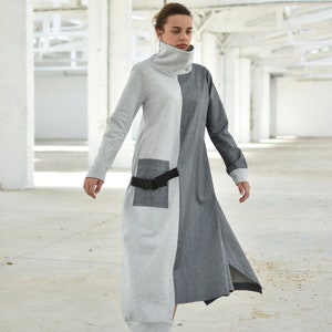 Puede incluir: Una mujer lleva un vestido largo, gris y gris oscuro con un cintur&oacute;n negro. El vestido tiene cuello alto y mangas largas. Lleva zapatillas blancas.