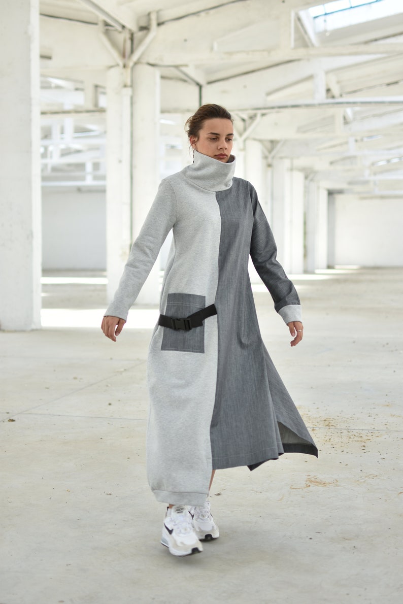 Puede incluir: Una mujer lleva un vestido largo, gris y gris oscuro con un cintur&oacute;n negro. El vestido tiene cuello alto y mangas largas. Lleva zapatillas blancas.