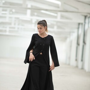 May include: A woman wearing a black long-sleeved dress with a split in the front and a button closure. The dress has wide sleeves with slits at the wrists. She is standing in a white room with a white ceiling.
