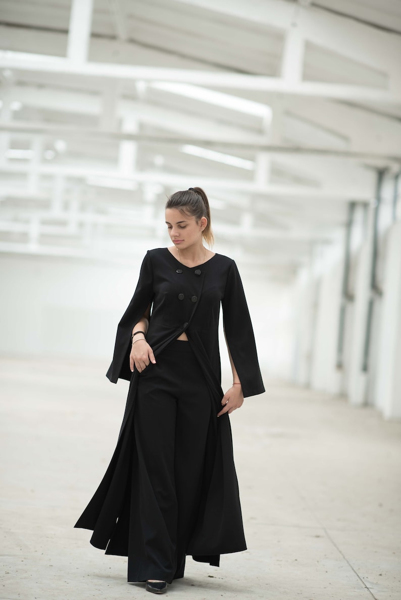 May include: A woman wearing a black long-sleeved dress with a split in the front and a button closure. The dress has wide sleeves with slits at the wrists. She is standing in a white room with a white ceiling.