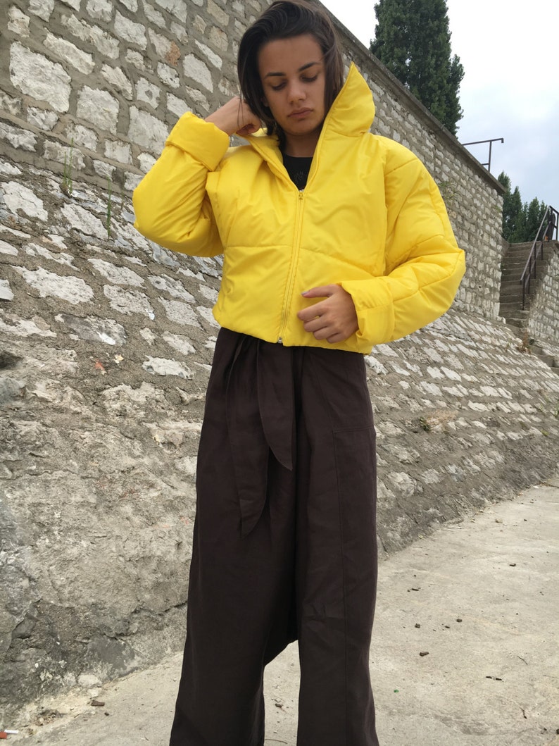 May include: A cropped, bright yellow puffer jacket with a high collar and a front zipper. The jacket is paired with dark brown, wide-leg pants. The outfit is worn outdoors against a stone wall background.