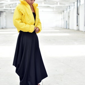 May include: A woman wearing a bright yellow cropped puffer jacket and a long black skirt. The jacket has a high collar and a quilted texture. The skirt is flowing and asymmetrical, creating a modern silhouette. The outfit is set against a white background.