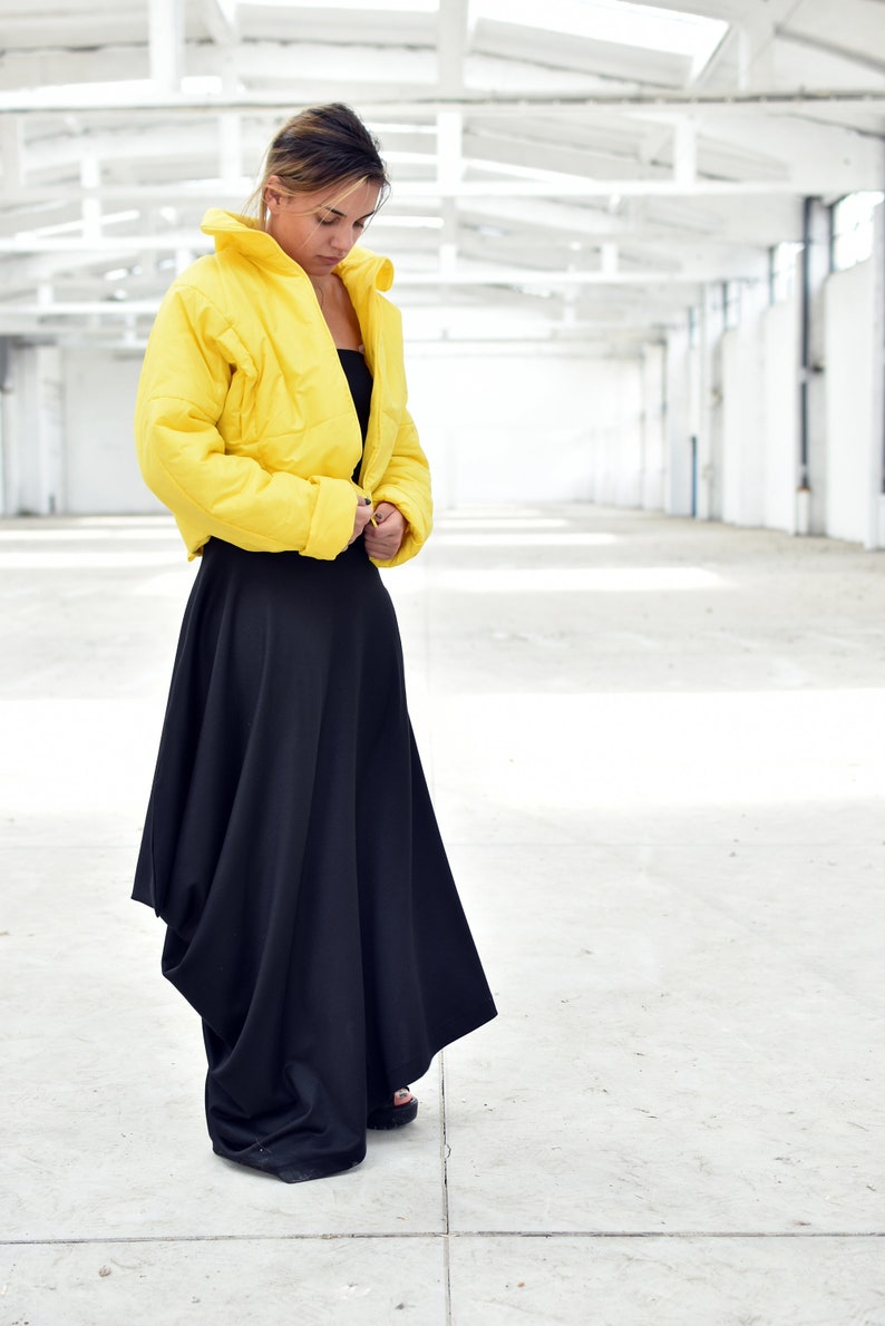 May include: A woman wearing a bright yellow cropped puffer jacket and a long black skirt. The jacket has a high collar and a quilted texture. The skirt is flowing and asymmetrical, creating a modern silhouette. The outfit is set against a white background.