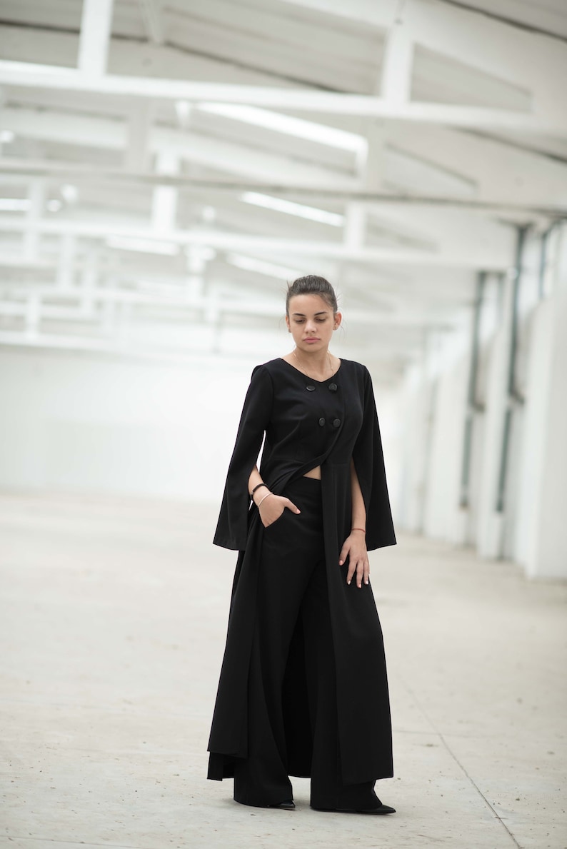May include: A woman wearing a black long-sleeved top with a high slit and wide-leg trousers. The top has a cape-like design and two buttons at the neckline. The woman is standing in a white, industrial-looking space.