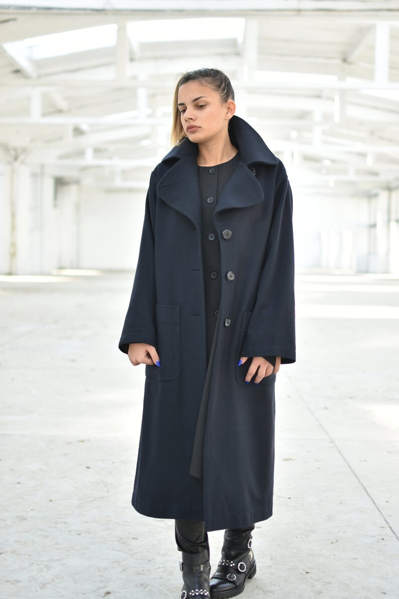 May include: A long navy blue coat with a large collar and two pockets. The coat has four buttons down the front and is worn over a black dress.