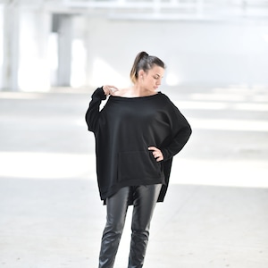 May include: A woman wearing a black oversized sweater with a pocket and black leather pants. She is standing in a white industrial setting.